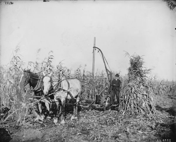 Man in a suit standing with a horse-drawn corn binder and a corn shocker in a field. A shock of corn is at his side.