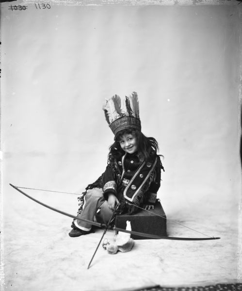 A young girl posing in Native American costume with bow and arrow in a studio. The image was likely used as a model for an advertising artist.