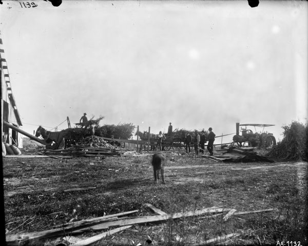 Group of men working with a tractor-powered husker and shredder in a farm yard. Men are standing on wagons, and there is a pile of boards in the yard. Part of a barn is on the far left. The blurred image of a dog is in the foreground.