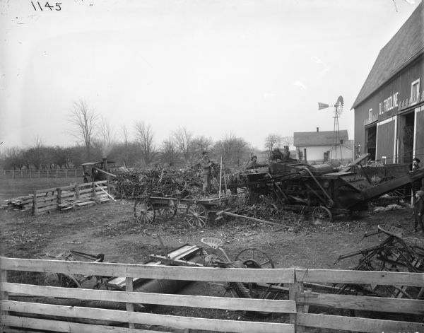 Men with a wagon feeding corn stalks into a husker-shredder near a barn. Other farm implements are in the foreground behind a fence, and a windmill is in the background.
