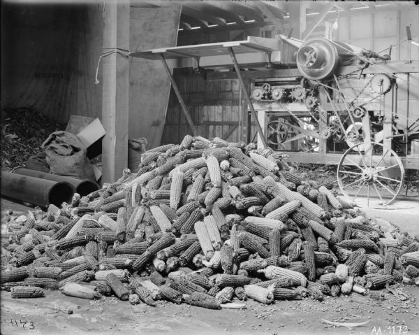 A pile of corn cobs in a large room, possibly a barn. Machinery with gears and belts is in the background.