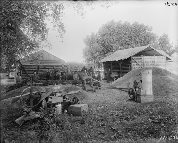 Several men and horses cutting ensilage(?) near farm buildings or sheds. The cutter is connected to a stationary engine with a belt. A pile of farm machinery parts and boxes is in the foreground on the left.
