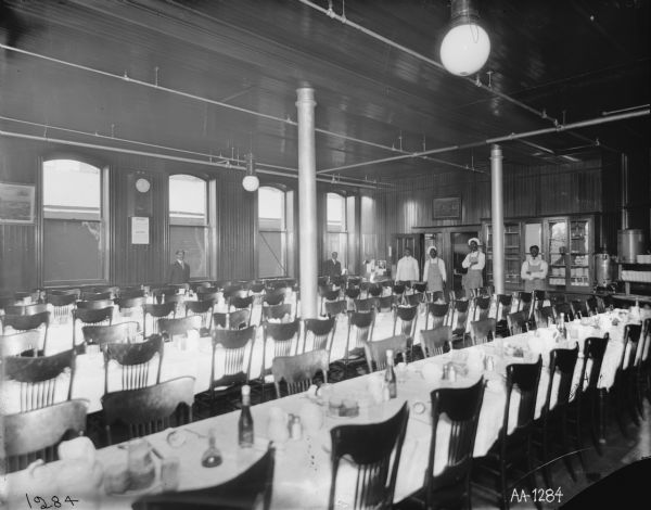 Long tables and chairs in a dining hall, probably at the McCormick Works. The tables are covered in table cloths and several cooks stand in the background.  On the wall there is a clock and a sign that reads, "NOTICE." Round light fixtures hang from the ceiling and cabinets are filled with dishes. Two framed advertising posters are on the walls: "Back from the War" and "Battle of Atlanta."