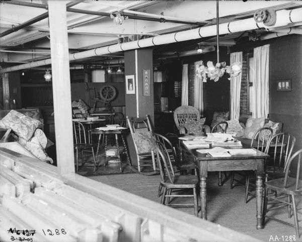 Chairs and tables are arranged throughout a room, possibly a factory lounge or break room, probably in the McCormick Reaper Works. A spinning wheel is in the background and books and magazines are arranged on the tables.