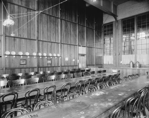 Factory dining room with a very high ceiling, probably at the McCormick Reaper Works. Long tables, stained with use, are lined with chairs. A row of decorative plates are on one of the walls. Curtains cover part of the windows.