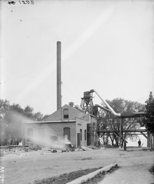 Men standing around a brick processing facility that may be near a body of water. A burn-barrel is in the foreground.