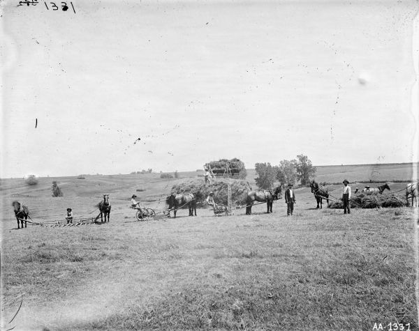 Men use horses, mowers, rakes and a hay loader to harvest hay in a field. A man is standing on a large haystack in the background.