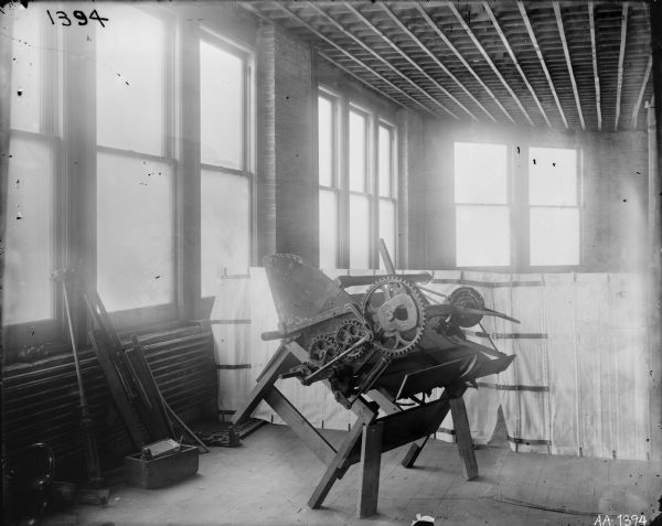 Piece of machinery, possibly part of a grain binder, propped up on a factory floor, most likely at McCormick Works. There is a white sheet hanging in the background for a backdrop.
