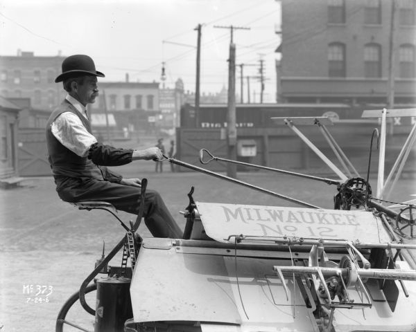 Right side view of a man sitting on a Milwaukee No. 12 Harvester and Binder (grain binder). In the background are men standing by a large metal gate and railroad cars. Neighborhoods with buildings are in the background. Likely taken on the grounds of the McCormick Works.