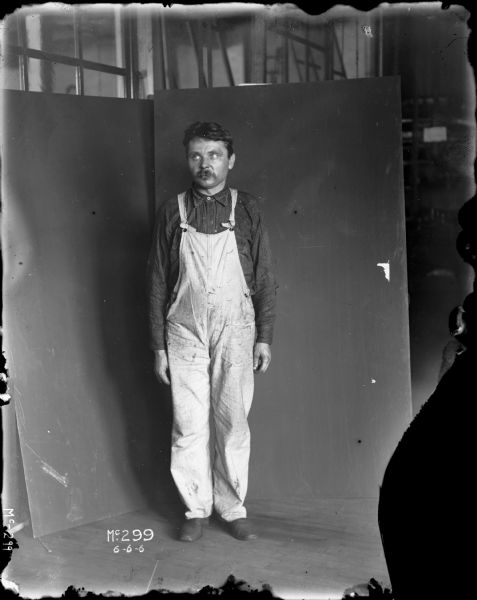 Full-length portrait of a man standing in front of a dark-colored makeshift background. He is wearing dark work shoes, light-colored overalls, and a dark striped button-up shirt buttoned to the collar. The man has a full dark moustache and short dark hair. His arms are to the side and he is looking to his right. The picture was likely taken at McCormick Works.