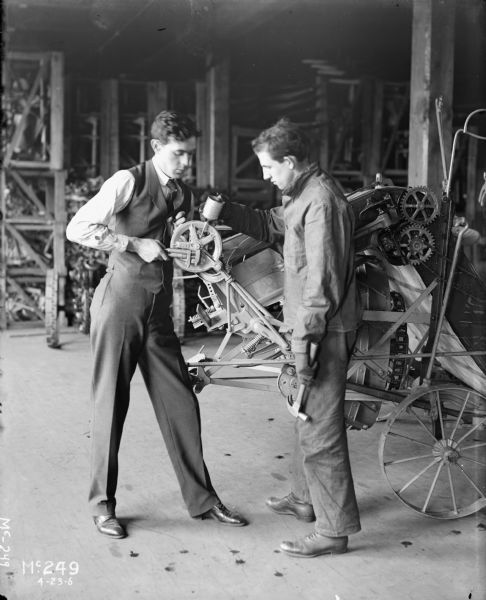 Two men standing on what appears to be a factory floor, probably the McCormick Works, oiling a wheel. The man on the left is dressed in suit pants, vest and tie, while the man on the right holding the oil can and a hammer is dressed in work clothes. The men appear to be working on a grain binder.
