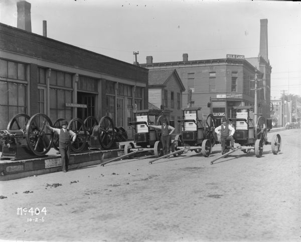 Three men are standing posing in street near a loading dock with early International Harvester stationary engines. The man on the left is standing with what appears to be three horizontal stationary engines, while the men on the right stand with vertical stationary engines in carts. All three men are wearing dark pants, vests, light-colored shirts, and dark hats. In the background are signs for a store selling milk cans. The loading dock also has the business name and advertisements on the curb.