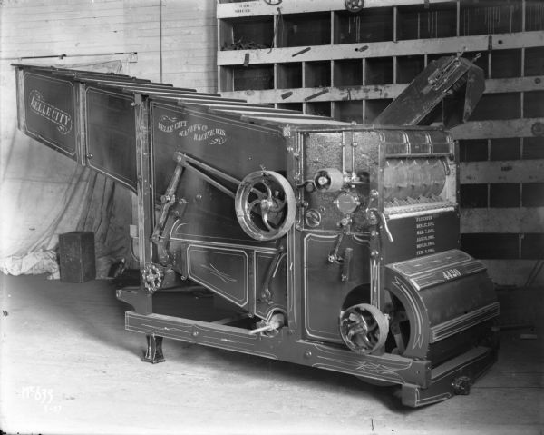 A Belle City Manufactory Co. thresher sits in what may be a factory, possibly the McCormick Works. In the background is what appears to be a supply shelf and a door blocked by a light-colored curtain.