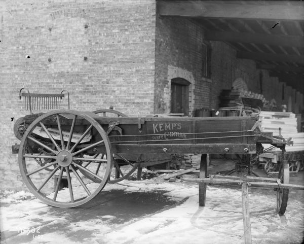 A Kemp's Manure Spreader is sits on a snow-covered loading dock of a brick building, possibly the McCormick Works factory. The side of the manure spreader reads: "Kemp's 20th Century". In the background lumber and other materials are stacked under the roof-covered area of the loading dock.