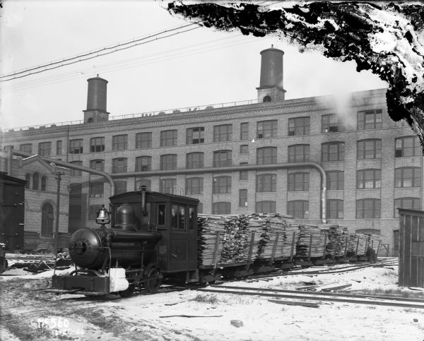 Six flat cars loaded with lumber are lined up behind a locomotive in a factory rail yard, probably at the McCormick Works. In the background is a large factory building and a few smaller buildings. A sign along the top of the building partially reads: "... WORKS."