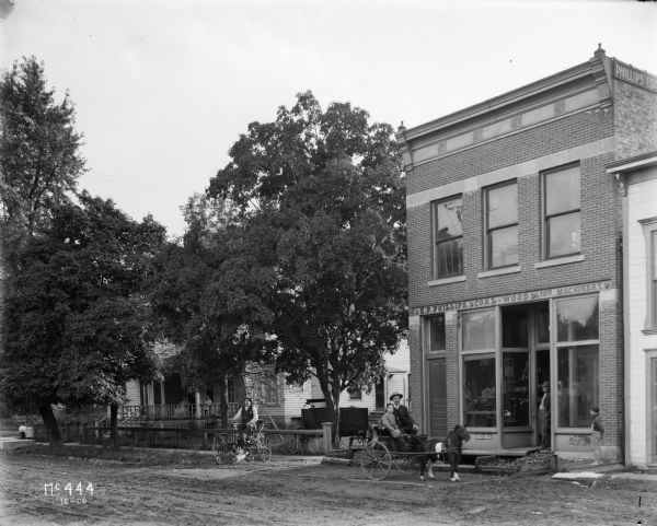 Two buggies are sitting parked in from of "E.P. Phillips Coal Wood & Machinery" store, possibly an International Harvester dealership. The first buggy is pulled by a miniature horse, and a boy and man are sitting in the buggy. On the left a man is sitting in a second buggy. The storefront is on the right, with a man leaning in the store doorway and a boy standing on the sidewalk nearby. In the background on the left is a large home with two women sitting on the front porch. On the extreme left two small children are standing on the sidewalk.