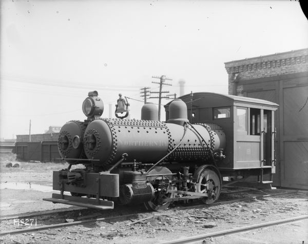 A locomotive for the Northern Railway is backed up near the closed doors of a brick building which sits on the railroad tracks, probably on the grounds of International Harvester's McCormick Works. The side of the locomotive reads: "Illinois Northern RY". On top of the locomotive is a large bell and on the front is a light. In the background there are fences, buildings, smokestacks and some power lines.