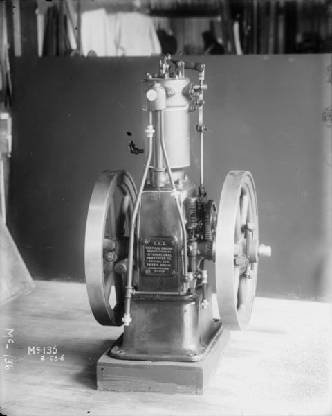 An IH vertical engine engine is sitting on a table with a dark background at McCormick Works. The plate on the front of the engine reads: "I.H.C. Vertical Engine Manufactured By International Harvester Co. Chicago, U.S.A. Patents Pending H.P. (?) Speed (?) No. 801." The numbers after horsepower and speed are illegible.