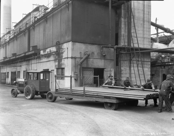 Five men loading pipes onto a flatbed trailer being pulled by an IH 10-20(?) industrial tractor. In the background is a large building, probably a manufacturing plant or warehouse.
