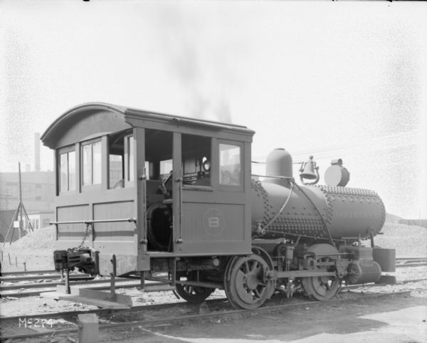 A locomotive for the Northern Railway sits on railroad tracks, probably on the grounds of the McCormick Works. The side of the locomotive reads: "Northern RY" and there is a large bell on top. The conductor's car has the number "8" painted on the side.