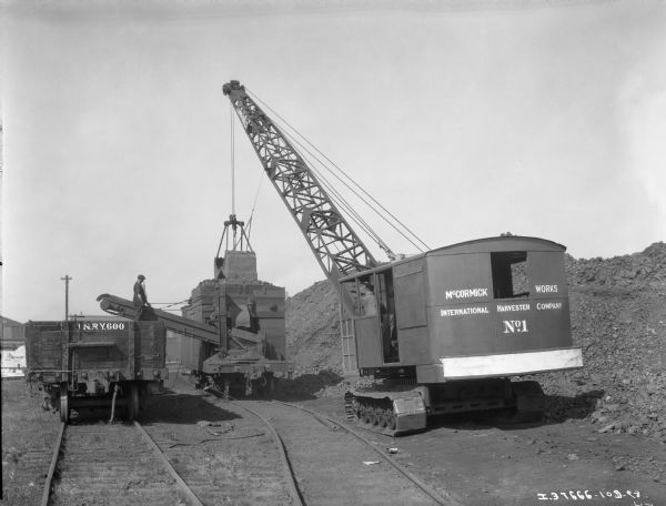 A man is sitting in the cab of a "McCormick Works International Harvester No.1" crawler-based crane with an earth moving attachment. The crane is loading a railroad car which has a conveyor belt, probably for sifting for natural resources, with dirt from a large pile in the background. A man is standing in another railroad car on the left where the conveyor belt empties.