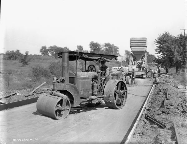 A man operating what appears to be a 10-20 Industrial tractor equipped with an Acme Road Roller(?) attachment. In the background a man is sitting in a vehicle. Six other men are assisting in the paving of a road. A cement truck is in the background.