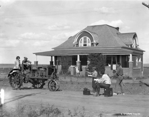 Cameramen staging a shot of a man and woman on a Farmall Regular tractor in front of farmhouse at an International Harvester demonstration farm in Gull Lake, Saskatchewan, Canada. The cameramen worked for International Harvester's Advertising Department.