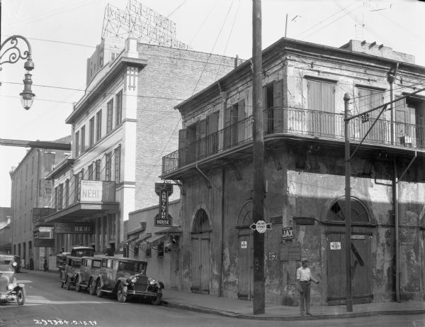 Street scene featuring the Old Absinthe House, a famous New Orleans establishment in the city's French Quarter. The building was built in 1806 by Pedro Front and Francisco Juncadelia of Barcelona to house their importing firm. In 1815, the ground floor was converted into a saloon known as "Aleix's Coffee House." It was renamed "The Absinthe Room" in 1874. A man is standing on the corner in front of the building with a tube under his arm. In the background is a building containing the offices of the American Brewing Company, bottlers and distributors of Nehi soft drinks.