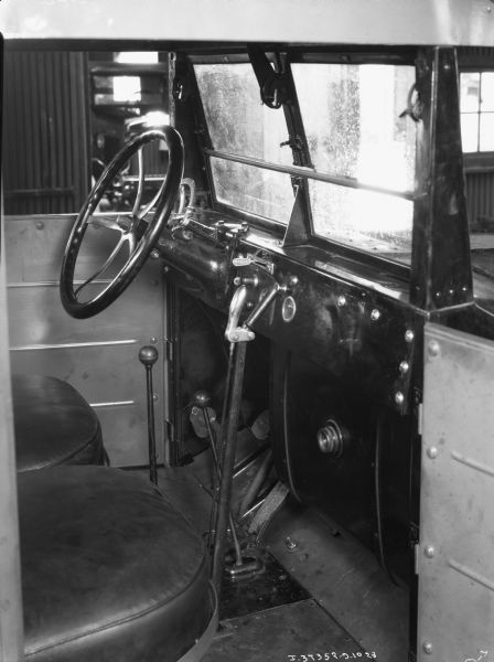An interior view of an International truck, showing the steering wheel, gear stick, column shifter, acceleration pedal, brake pedal, and clutch. There is an instrument panel on the dashboard.