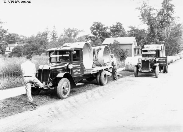 Two IH trucks for "W.E. Draper Cartage" are parked on the side of the road. Both trucks are hauling large concrete tubes, probably for construction work. The driver's side door of the trucks read: "W.E. Draper Cartage -Phone- (?)32.J" The text before the phone number is difficult to read, could possibly be a city starting with "T" in Florida.<p>One man is standing and bracing his foot on the front bumper of the car with his back to the camera, while a second man is working with the concrete tubes in the back of the truck. The second man is wearing work overalls and a hat while the first man is dressed more formally. Behind the trucks is a wooded area and a small wooden house. 
