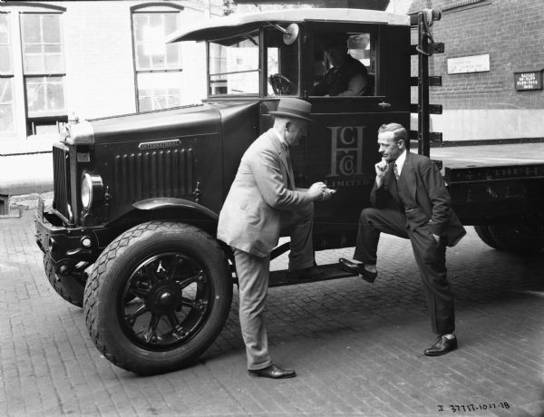 A man is sitting in an International delivery truck for the "H.C. Co. Limited." The "International" emblem is on the front side of the truck and the company name "H.C. Co. Limited" is on the driver's side door. The truck bed is empty, and the beginning of the company name "The Ha..." is on the bed. Two men in suits appear to be having a conversation next to the truck while leaning on the driver's side running board. There are brick buildings in the background, and a sign to the right reads: "Danger Go Slow, Blow Your Horn" next to another sign that says: "Shipping and Receiving Dept."