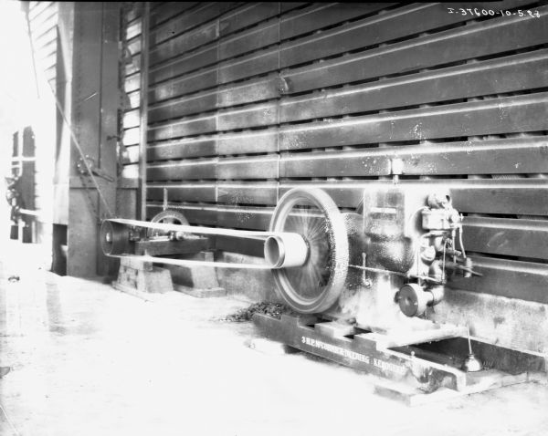 View of a stationary McCormick-Deering kerosene engine along a wall. On the side of the engine is written: "3 H.P. McCormick-Deering Kerosene." To the right of the engine is an oil can.