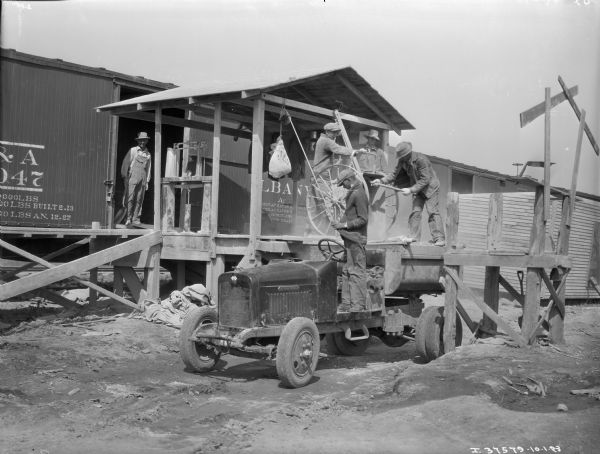 One man is standing on an IH industrial tractor driver's seat while three other men are loading what appears to be a farm implement onto a trailer. The "International" emblem is on the front side of the tractor. The men are standing on a railroad dock, unloading the implement directly from the railroad car. There is a fifth man standing in the open doorway of the railroad car.