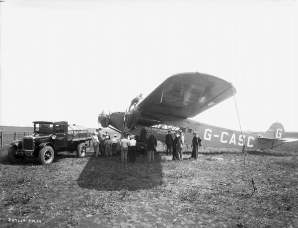 A group of fifteen men and boys are standing by while an International model SF-46 gasoline truck fuels a Western Canada Airways tri-motored airplane. One man is standing on top of the airplane. The photograph was taken in Edmonton, Alberta, Canada.