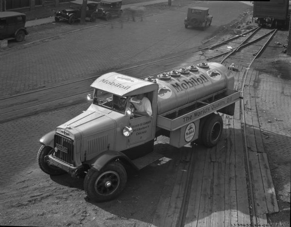 Elevated view of a man sitting in the driver's seat of an International delivery truck for the "Vacuum Oil Company." The top of the cab reads: "Mobiloil" while the driver side door reads: "Vacuum Oil Company 343 S. Dearborn St. Chicago 8." The oil tank itself reads "Mobiloil" with logos on either side. The phrase "The World's Quality Oil" is on the side of the carrier with the "Mobiloil" logo beneath. The truck is parked on railroad tracks and in the background is a rail car, three parked vehicles on the grass, and one vehicle parked on the cobblestone street. There are blurred images of the figures of people near the parked vehicles.