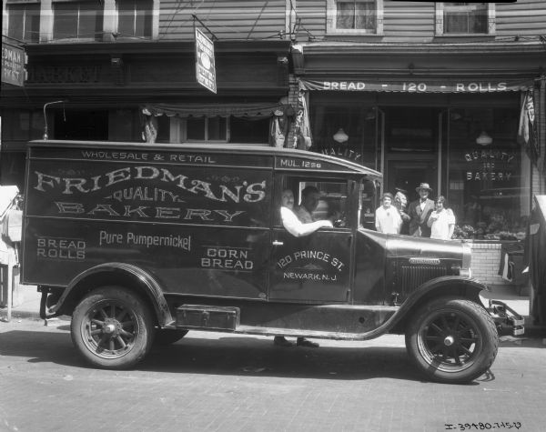 View across street towards two men sitting in an International delivery truck. Behind them standing on the sidewalk are a man holding a loaf of braided bread, and two young women. The side of the truck reads: "Wholesale & Retail Friedman's Quality Bakery, Bread, Rolls, Pure Pumpernickel, Corn Bread." The address on the truck reads: "120 Prince St. Newark, N.J." Behind the group is the storefront for the bakery, with signs on the windows reading: "Quality 120 Bakery," and an awning reading: "Bread - 120 - Rolls." There are baked goods on display in the large window, and there are signs overhead for other businesses.
