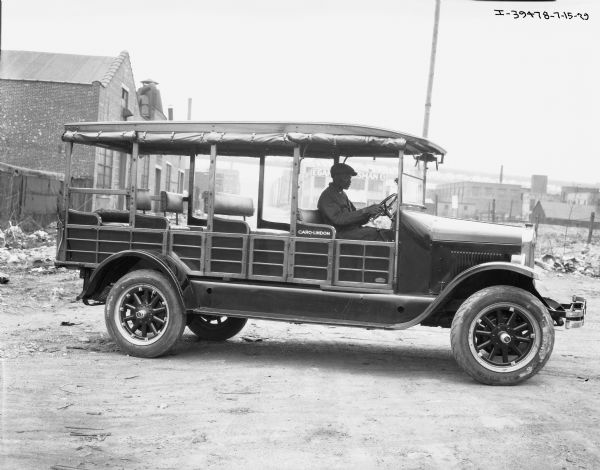 Side view of an African American man sitting behind the wheel of an International bus. The words "caro-lindom" appear on the side of the bus. In the background are industrial buildings.