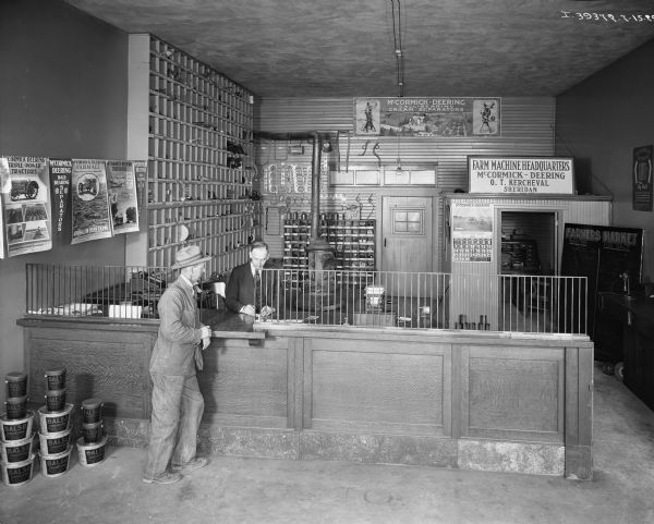 Elevated view of a customer at the sales counter of O.T. Kercheval's International Harvester dealership. Advertising posters and a calender are on the wall.