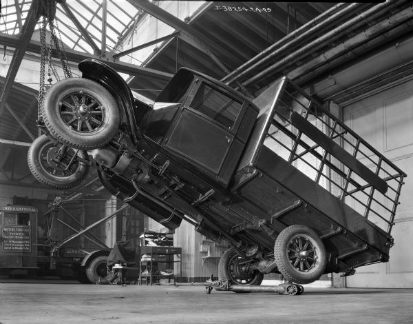 An International truck is hoisted at an angle in the air by chains in a service shop, most likely at an International Harvester dealership. In the background a mechanic's station is set up and and a tow truck is parked.