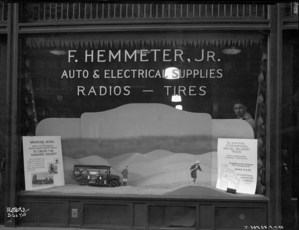A display window for the "F. HEMMETER, JR. AUTO & ELECTRICAL SUPPLIES RADIOS - TIRES" showing a scale model diorama of an International truck and two men in the sand dunes of the Sahara desert. A sign to the right reads: "MINIATURE MODEL OF THE FIRST FOUR WHEELED STOCK MODEL MOTOR VEHICLE TO CROSS THE SAHARA DESSERT On a trip over from Nairobi, British East Africa to London, England via Stanleyville Kano Algiers Marseilles and Paris." A sign on the left reads: "The identical International Harvester Truck Which preformed this remarkable achievement On display at our showroom 2565 Pennsylvania Avenue January 21 to 26 International Harvester Company." The store may have been located in Baltimore, Maryland. A man can be seen inside the window looking out at the camera.