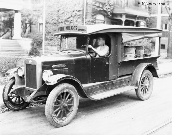 Side view of an International milk truck operated by "The Pure Milk Co. Limited." A man is sitting in the driver's seat holding the steering wheel. The back of the truck is loaded with milk cans and features a rolled up curtain. The "International" emblem is on the front side of the truck. In the background are houses.