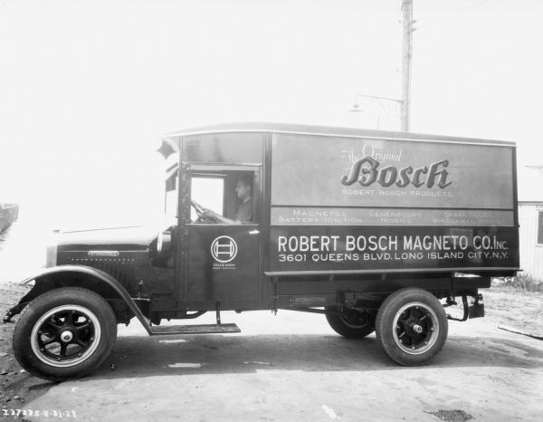 A man is sitting in the driver's seat of an International fleet truck for the "Robert Bosch Magneto Co." The vehicle appears to be parked in front of a body of water.
