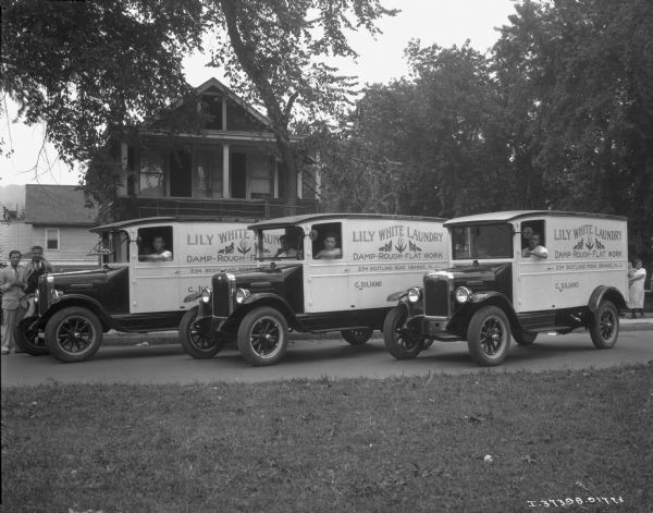 A fleet of three "Lily White Laundry" International trucks are staggered at an angle in the street. There is a driver in the driver's seat of each truck, and a passenger in the middle car. Signs painted on the side of the trucks read: "Lily White Laundry," "Damp-Rough-Flat Work," "234 Scotland Road Orange, N.J.," and "C. Juliano." The "International" emblem is on the front side of the trucks. Houses are in the background. On the left, standing in the street, are two men. On the far right a woman holding an infant is standing on the sidewalk.
