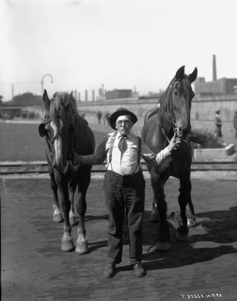 A man is standing between two horses, holding them by their bridles. The man is dressed in dusty shoes, dark pants with suspenders, a light shirt, a dark tie tucked into his shirt, glasses, and a dark hat. In the background are railroad tracks, an open field with men milling around, and possibly a cityscape. The man and horses may be at one of the McCormick-International factories.