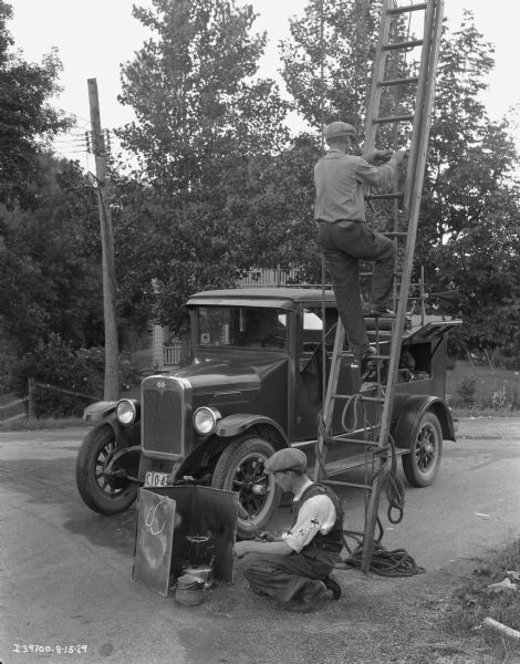 One man is climbing a ladder, while another man is crouched examining what appears to be a stove surrounded by a metal windbreak. In the background is an International truck with several open compartments filled with tools. There is a house in the far background behind trees.