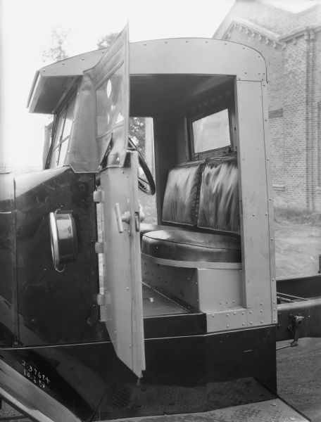 View through open driver's side door of an International truck cab, including seats and steering wheel. Behind the truck is a large brick building.
