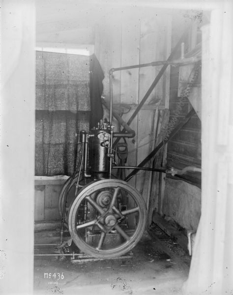 A stationary engine is sitting in the corner of what appears to be a shed. There are a number of pipes and wires attached to the engine. In the background, a window is covered with a dark piece of fabric.