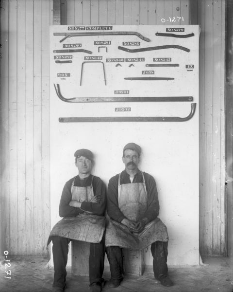 Two factory workers dressed in dark work clothes, light aprons, and dark hats sit on a crate below a numbered display of parts, most likely at International Harvester's Osborne Works. The display was probably assembled to aid in the creation of an owner's manual or parts catalog.