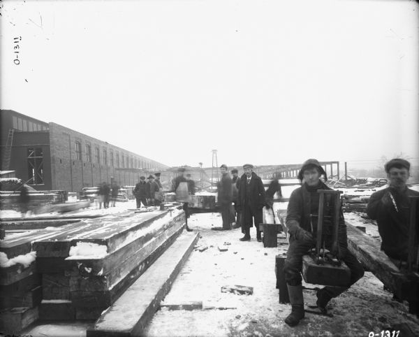 Several men are standing and sitting around a construction site at International Harvester's Osborne Works. Lumber is stacked in the yard, and in the background is construction equipment. The skeleton of a factory building is in the background behind the men. There is snow on the ground and the men are dressed in winter clothing. A couple of men are wearing aprons and one man is wearing an overcoat.