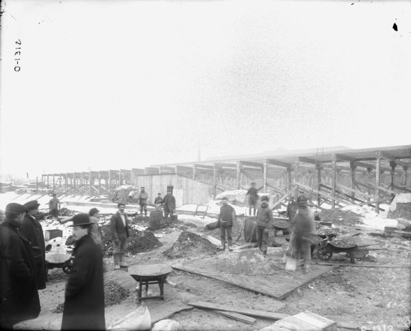 Several men are standing around a construction site at International Harvester's Osborne Works. There are wheelbarrows filled with building materials and some scattered lumber. The skeleton of a factory building is behind the men in the background. There is snow on the ground and the men are dressed in winter clothing. Three man are wearing overcoats and hats.